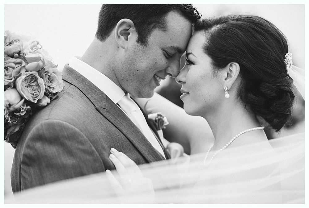 Bride and groom touching foreheads, smiling, close-up, black and white, outdoors.