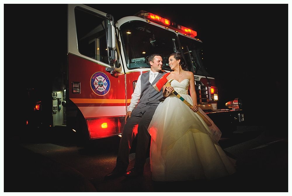Newlyweds pose in front of a fire truck at night; bride in white gown, groom in gray suit.