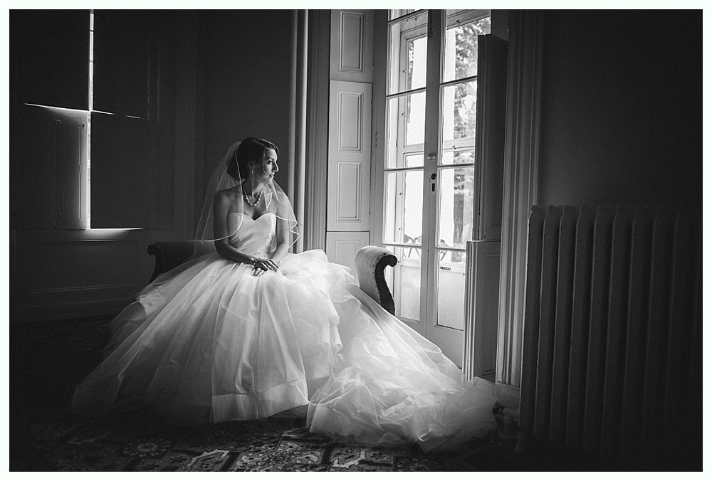 Bride in wedding dress sits near window, looking away. Black and white photo.