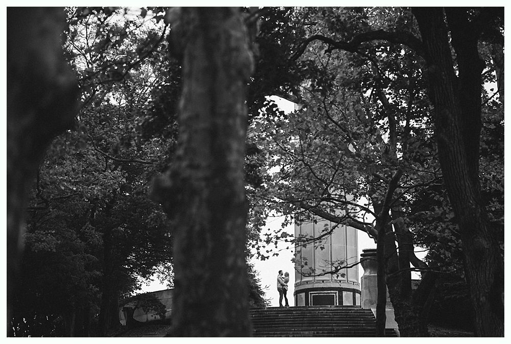 Couple standing at a monument, framed by trees. Black and white photo.