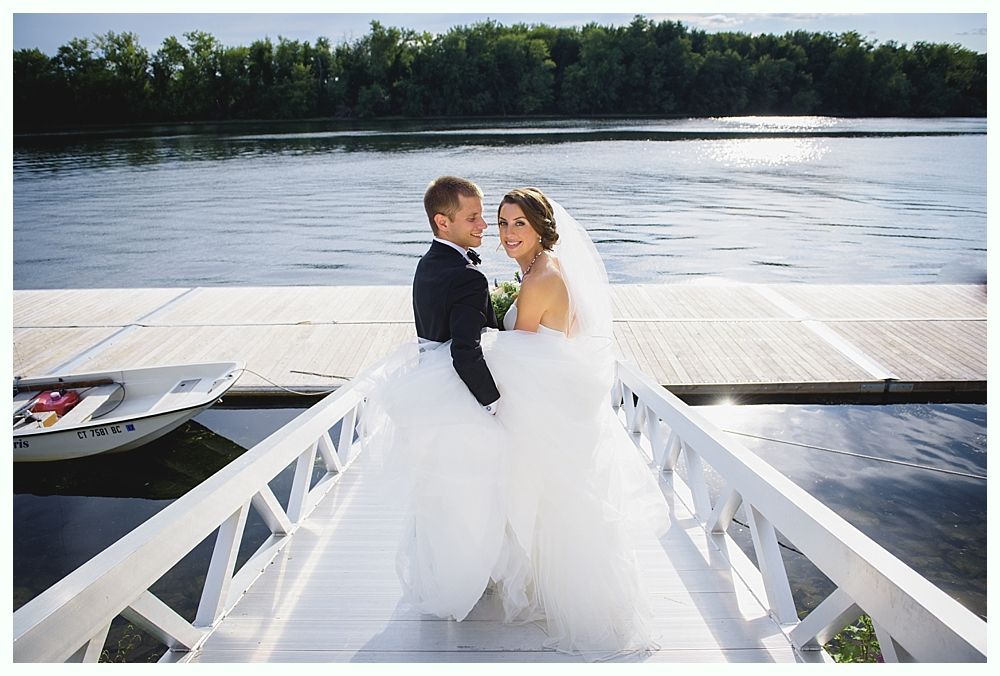 Bride and groom on a white dock, looking at camera. Water and trees in the background.