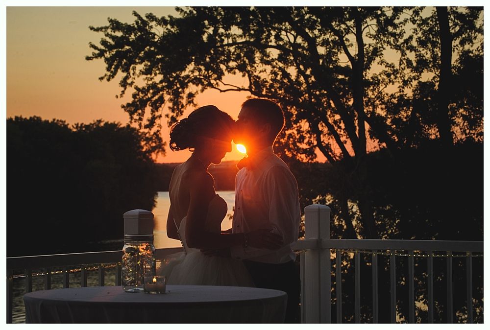 Couple kissing at sunset on a deck, silhouetted with glowing sky and water.