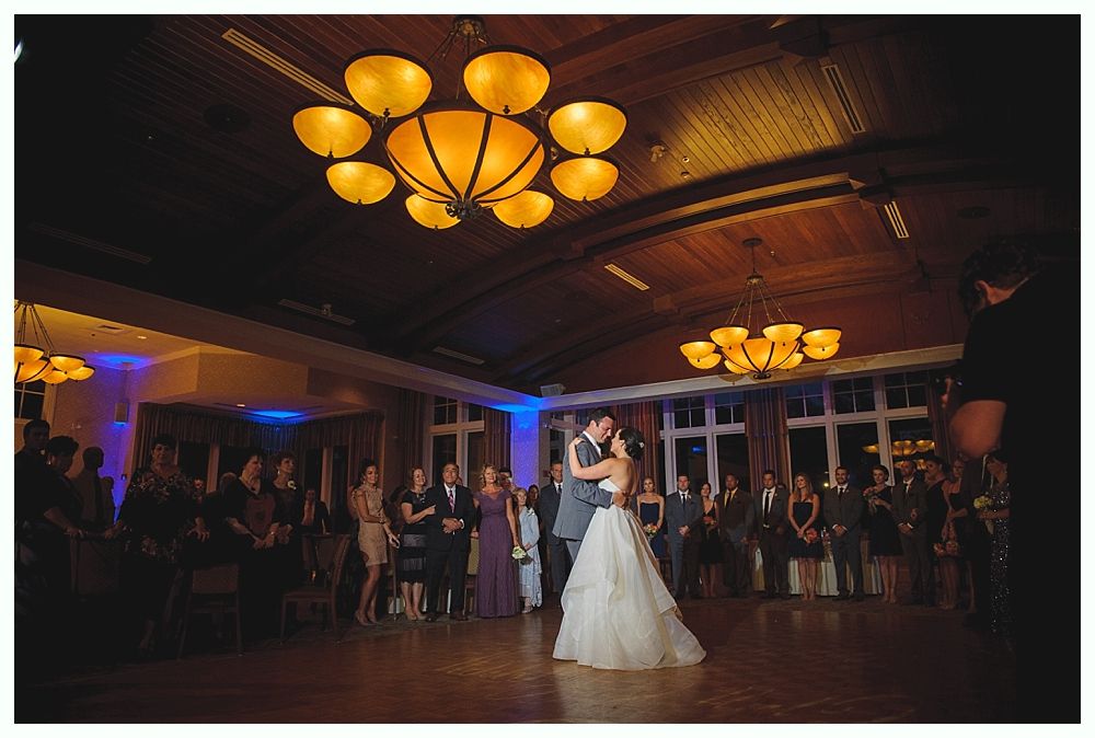 Bride and groom share a kiss on a dance floor, guests watch. Large chandeliers illuminate the space.