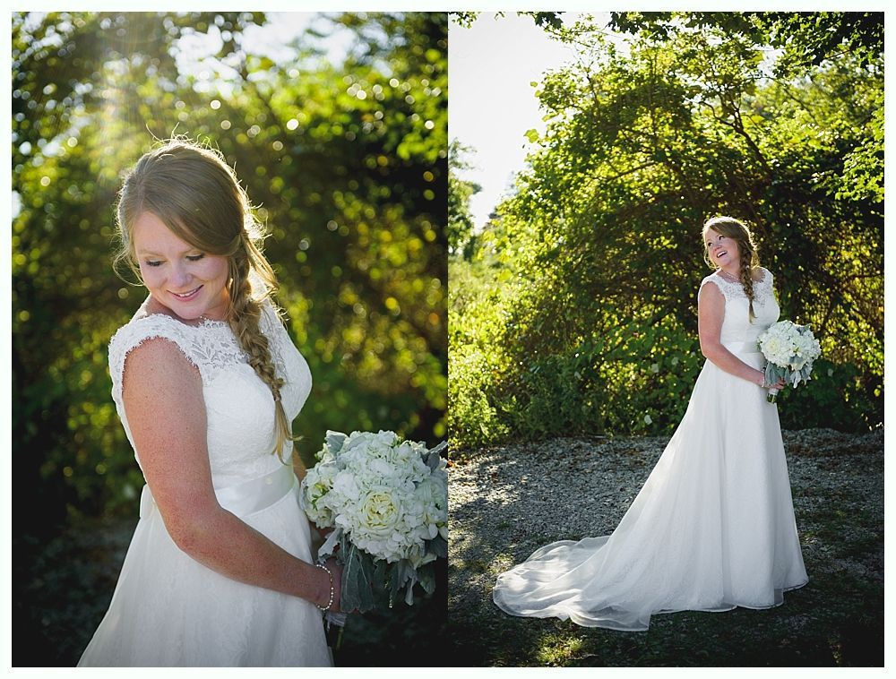 Bride in a white gown holds bouquet, smiling outdoors. Sunlight shines through the trees.