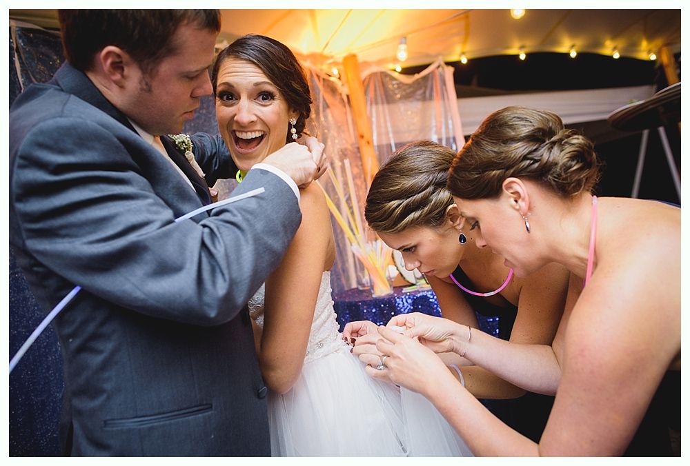 Bride smiles as friends and a man in a suit adjust her dress and veil; outdoor nighttime setting with string lights.