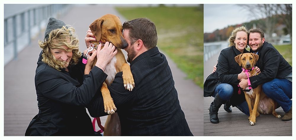 Two people cuddle a brown dog on a boardwalk. One person is kissing the dog. The people are smiling.