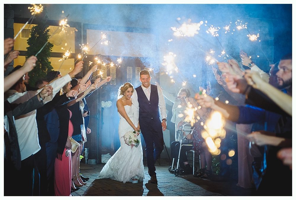 Couple walks through sparkler-lit pathway. Wedding guests hold sparklers; festive, nighttime outdoor setting.
