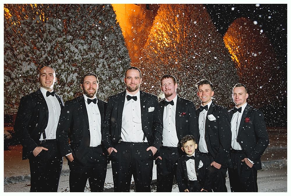 Men in tuxedos pose outside in the snow.