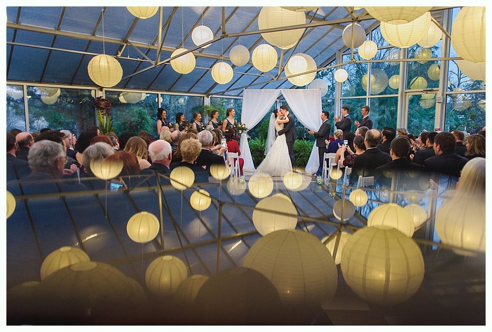 Wedding ceremony in a glass-enclosed space, decorated with hanging lanterns, reflected on the floor.