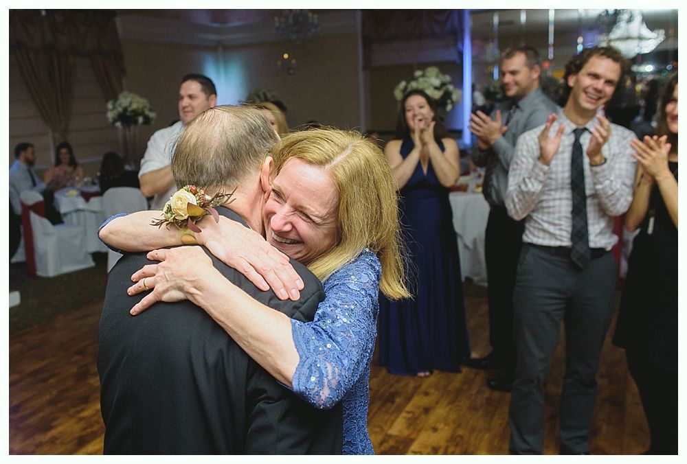 Woman embraces a man on a dance floor, as guests clap and watch during a celebration.