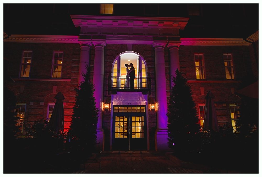 Couple embracing in a lit window of a building at night, accented with purple and red lights.