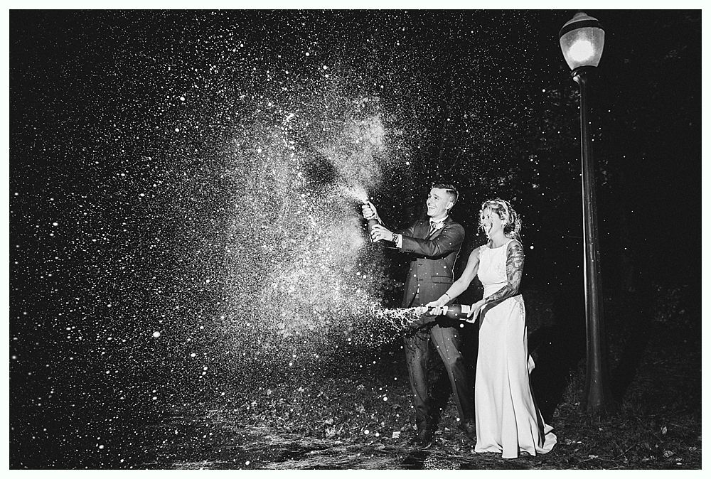 Couple popping champagne, spraying into the night. Lamp post illuminated on the right. Black and white photograph.