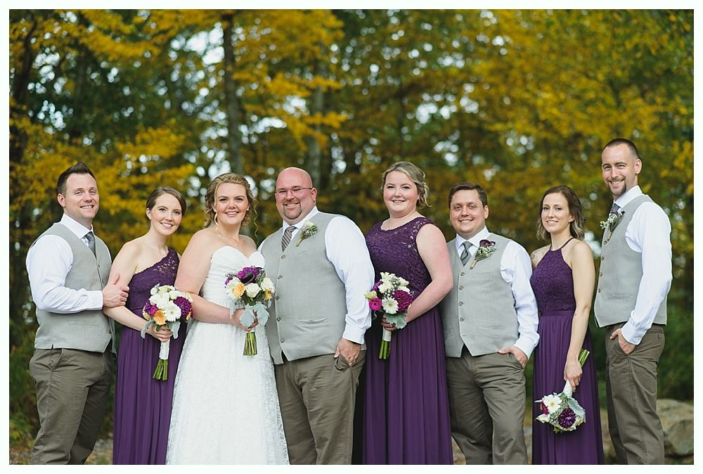 Wedding party posing outdoors; bride and groom centered with bridesmaids in purple and groomsmen in vests.