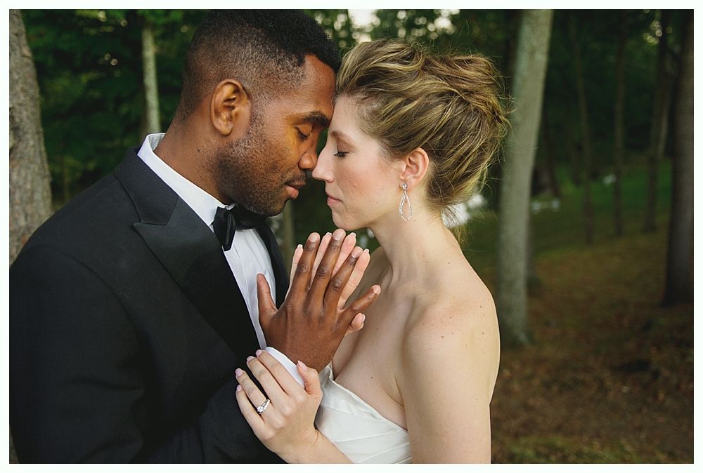 Couple embracing outdoors; Black man in tux, white woman in wedding dress, touching hands.