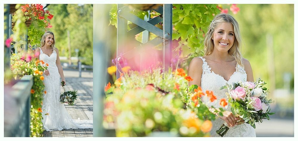 Bride in white dress holding flowers, standing near flowers and green foliage. Smiling.