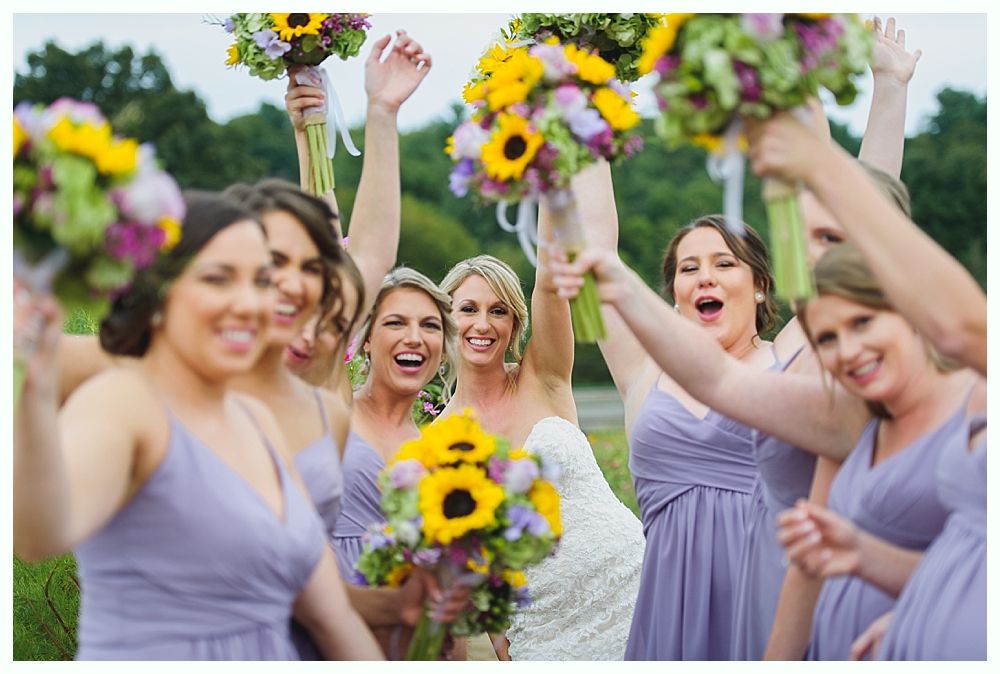 Bride and bridesmaids in lavender dresses with sunflower bouquets, celebrating outdoors.