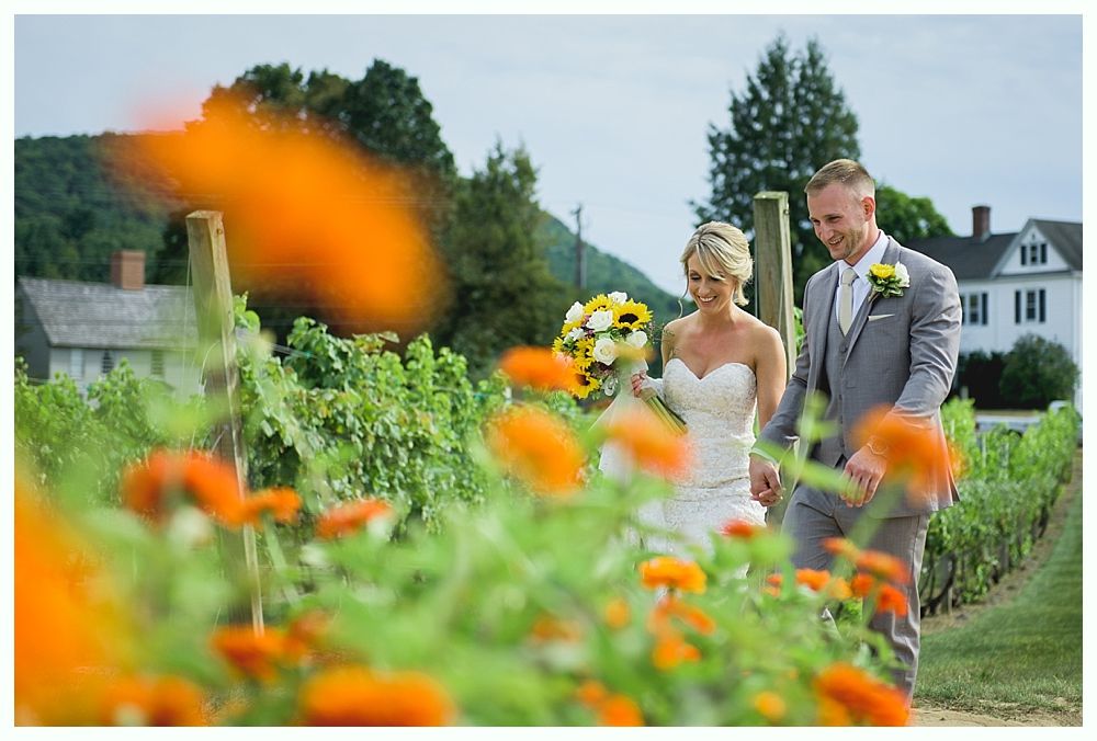 Bride and groom walking in a garden; flowers in foreground, holding hands, smiling. White house and hills in background.