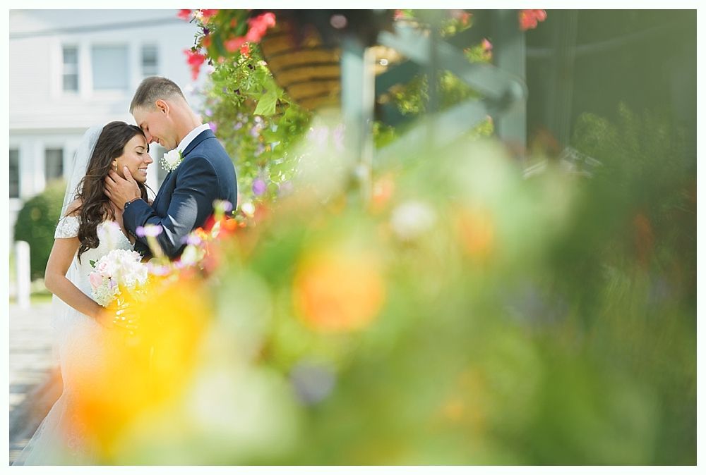 Bride and groom embrace by flowers; the bride holds a bouquet, the groom touches her forehead; a building is in the background.