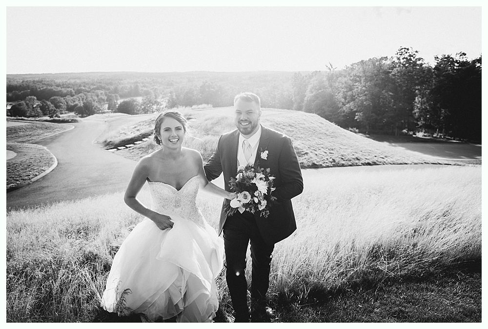 Bride and groom smiling, walking down a grassy hill after a ceremony. The scenic background includes a golf course and trees.