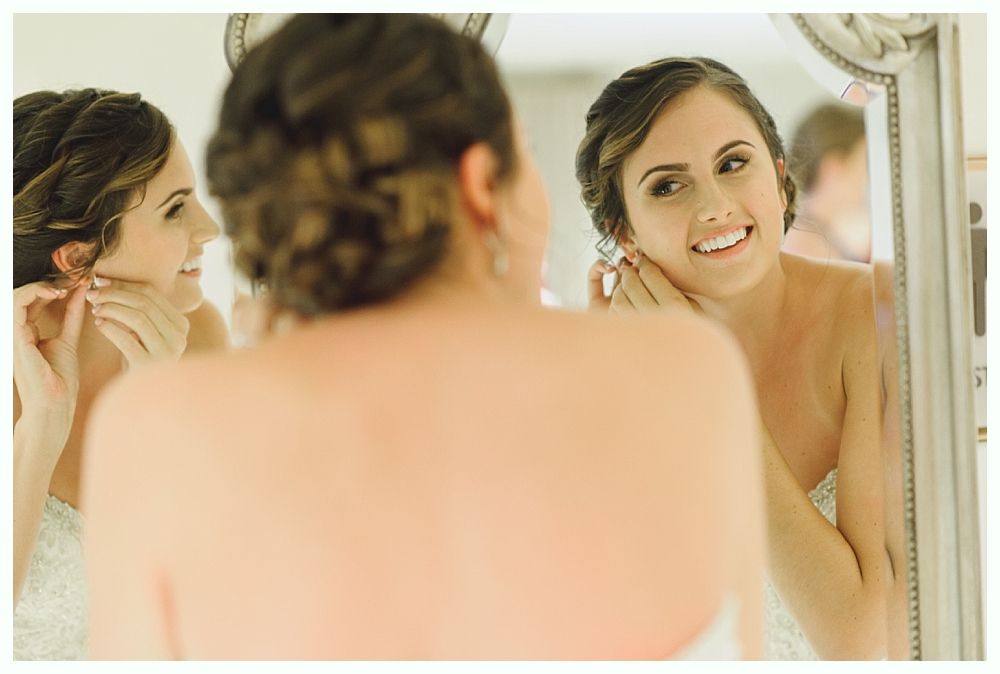 Woman in a white dress, smiling in front of a mirror, putting on earrings.