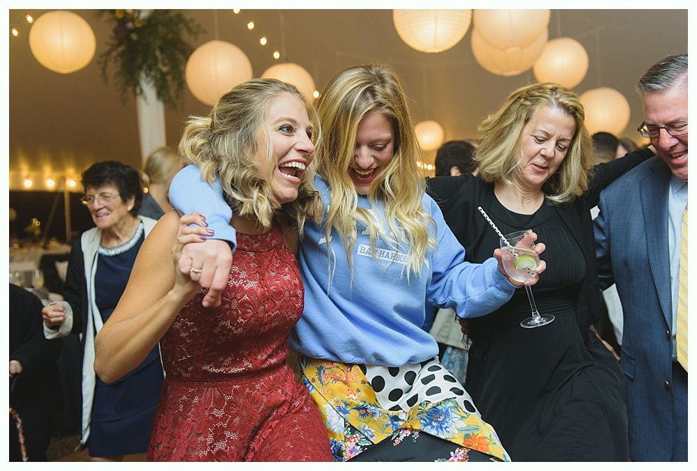 Three smiling women dancing at a party under hanging lights, one in a red dress, one in a blue sweatshirt.