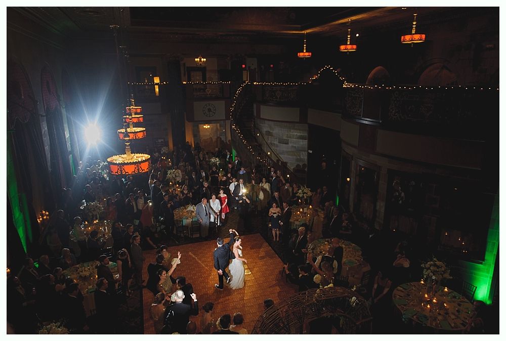 Wedding reception with couple dancing on a wooden floor, surrounded by guests at tables. Dim lighting with decorative chandeliers.