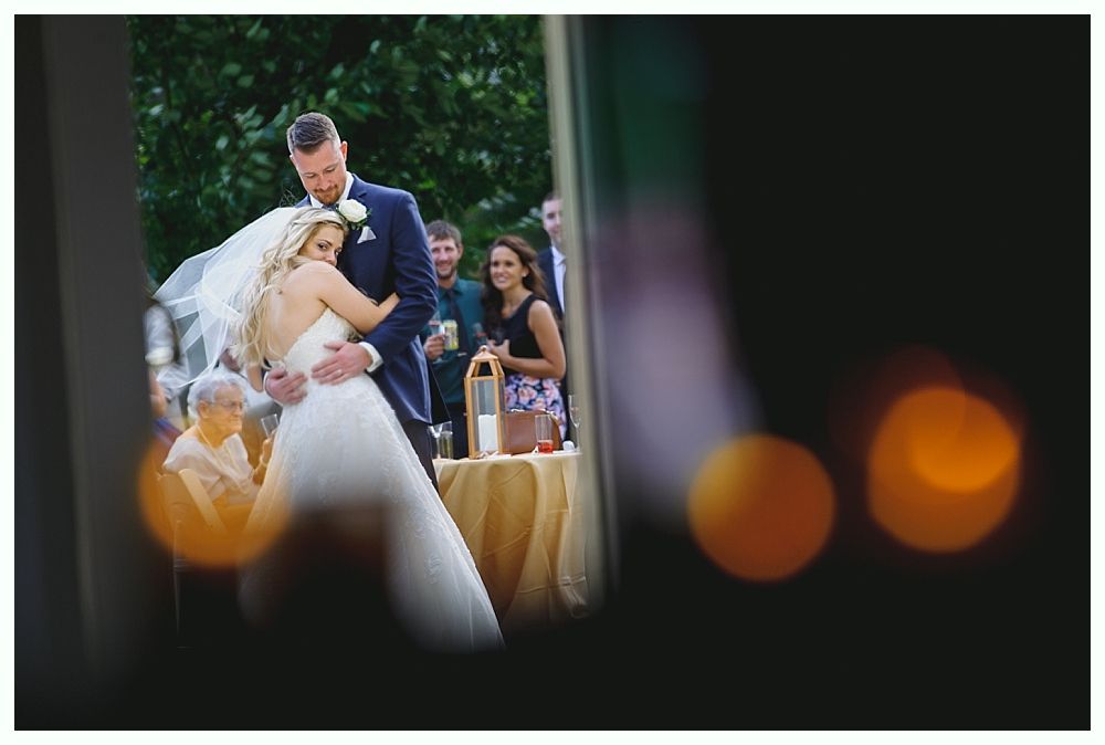 Wedding couple embraces; bride in white gown, groom in navy suit. Ceremony with guests outdoors.