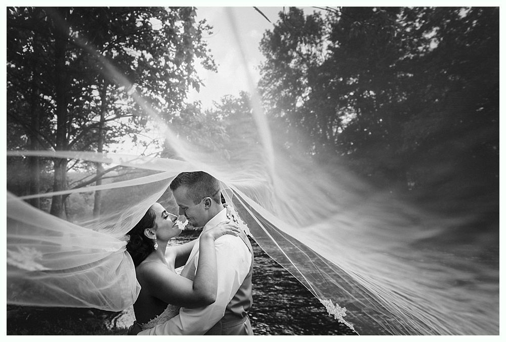 Couple embraces under a flowing sheer fabric, outdoors in front of a tree-lined backdrop, in black and white.