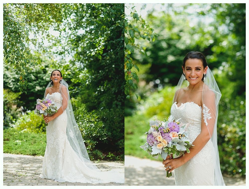 Bride in strapless lace gown holding bouquet, smiling outdoors with veil and greenery in background.
