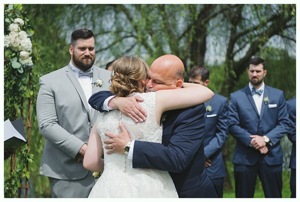 Bride hugs her father at outdoor wedding ceremony. Groomsmen watch.