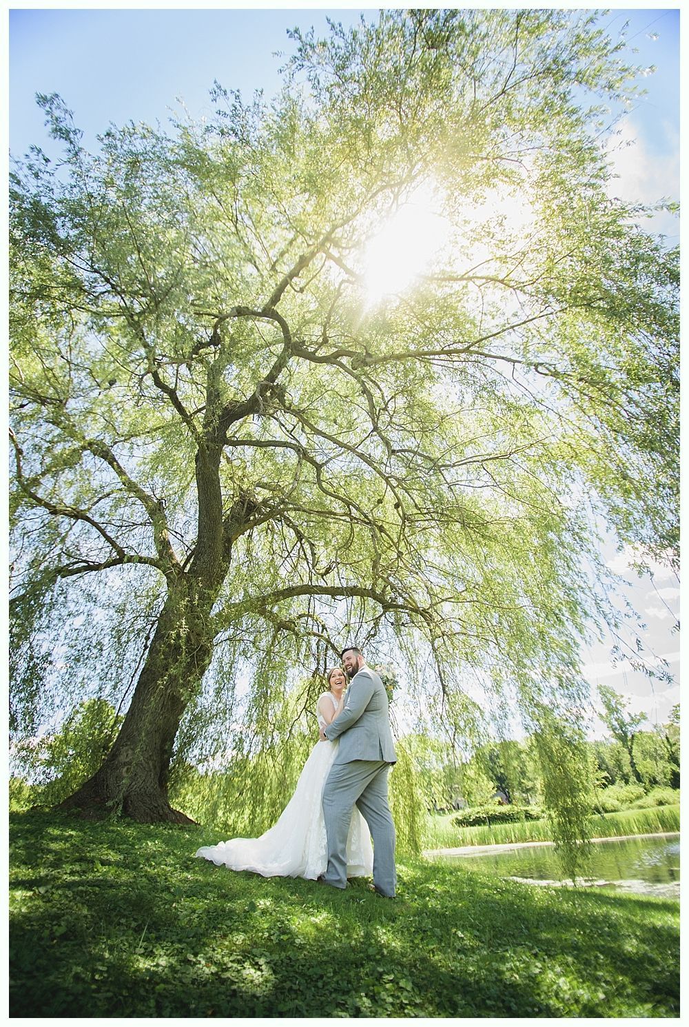 Bride and groom embrace beneath a willow tree on a sunny day near a pond.
