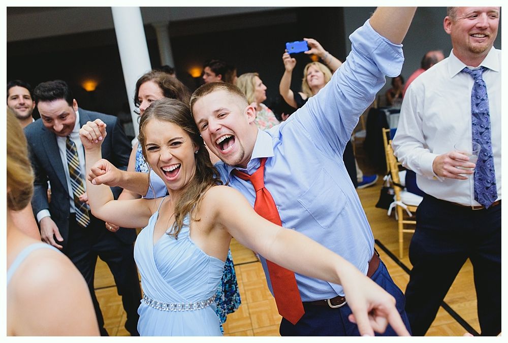 People dancing joyfully at an event; woman in blue dress, man with red tie, others in the background.