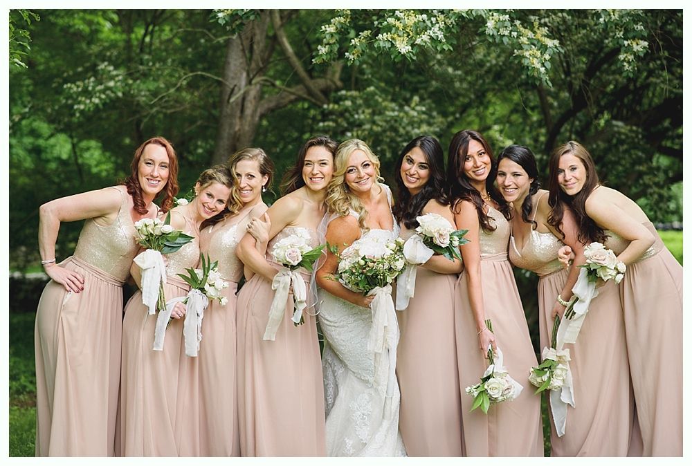 Bridesmaids in blush dresses pose with bride in white dress, holding bouquets, in a wooded area.
