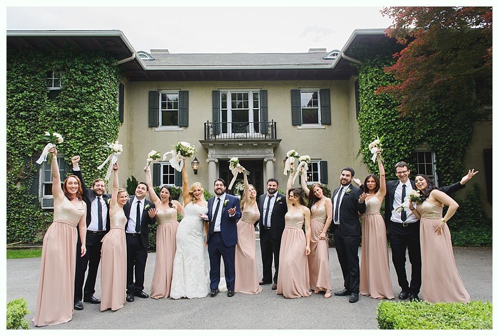 Wedding party in front of a large house, raising bouquets. Bridesmaids in pink, groomsmen in black suits.