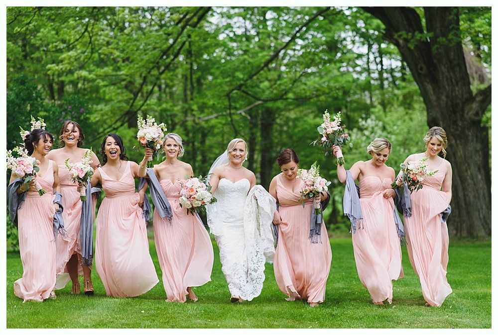 Bridesmaids in pink dresses and a bride in white laughing and walking in a grassy area with trees.
