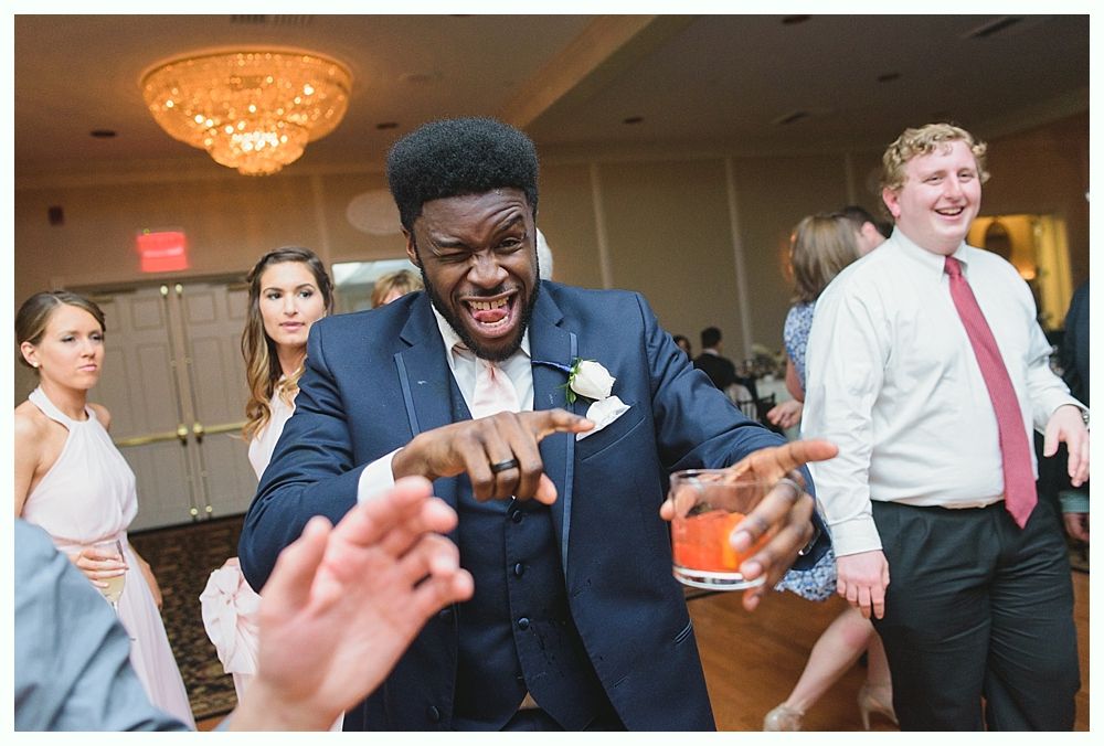 Man in suit winks, points and holds a drink while dancing at a wedding reception; others look on.