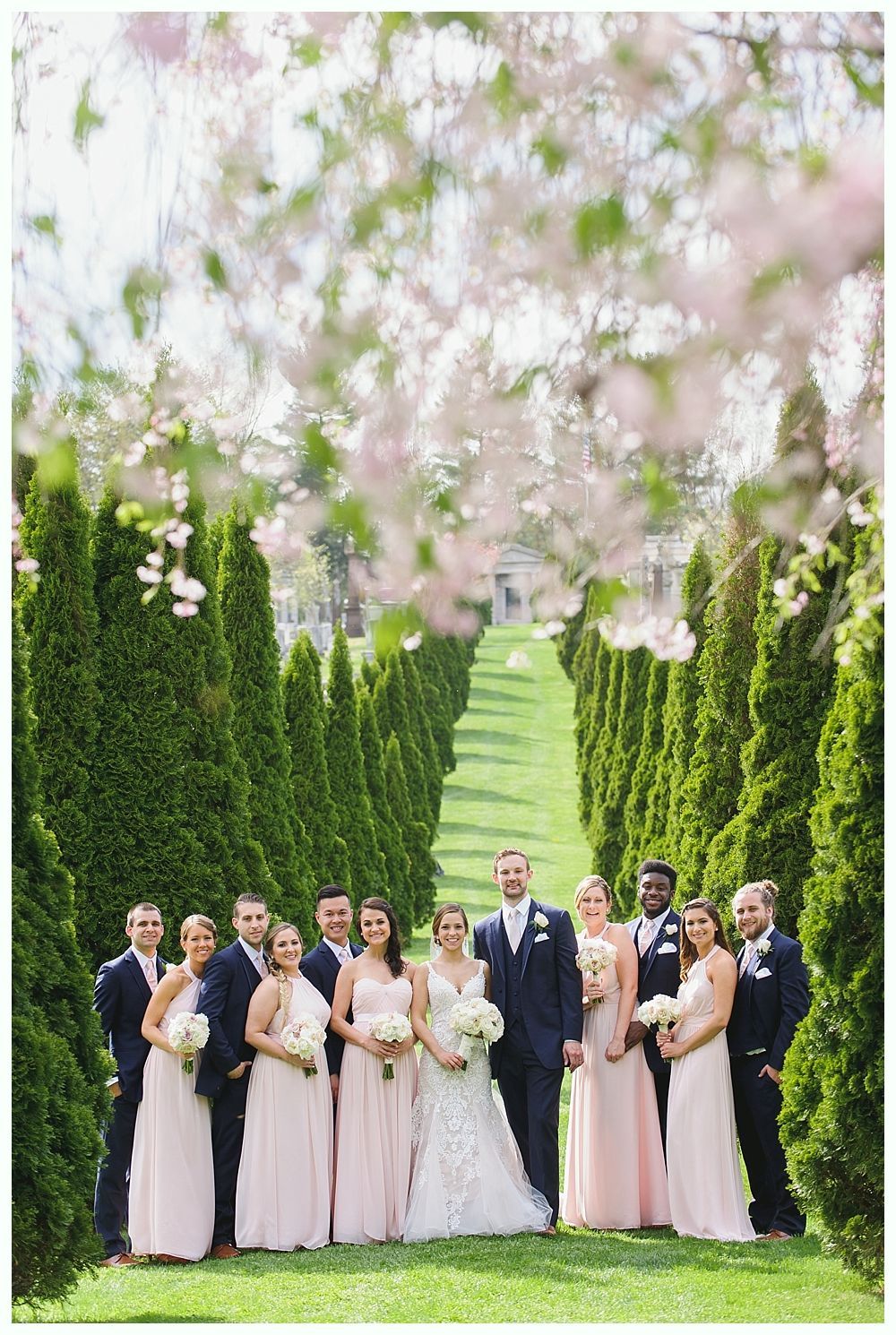 Wedding party posing on a green lawn lined with tall, green trees. Bride in white dress, bridesmaids in pink. Groom and groomsmen in navy.