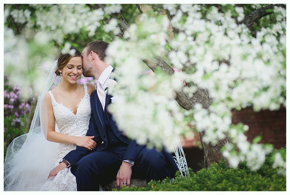 Bride and groom in a garden setting, she in a white dress, he in a blue suit, he kisses her cheek.