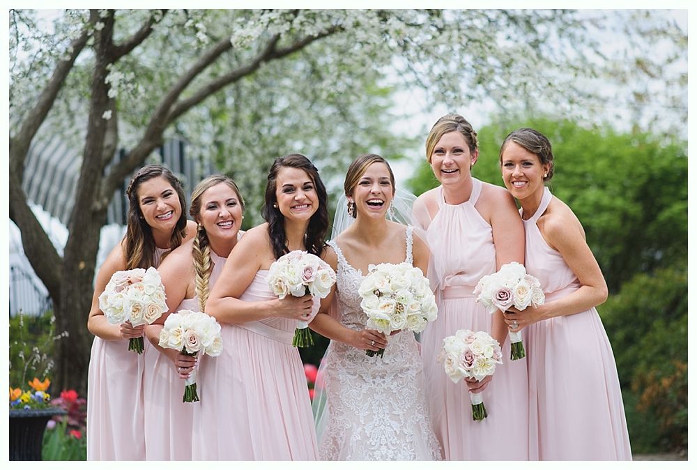 Bridesmaids and bride pose outdoors; all in pink dresses holding bouquets, smiling.