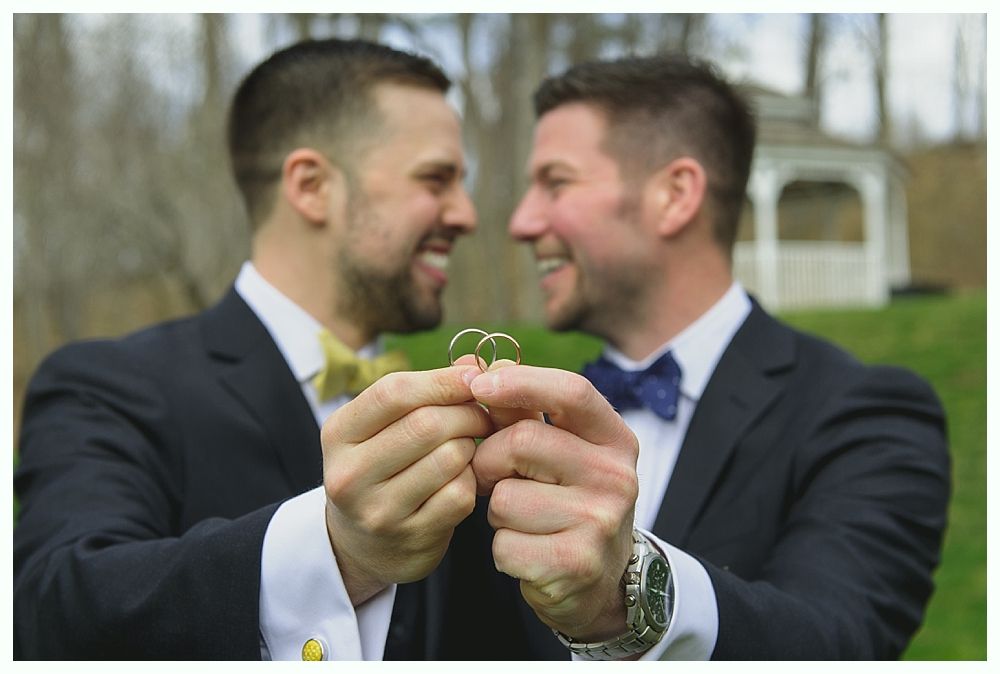 Two men smiling, holding up wedding rings. They are wearing suits with bow ties, outdoors.
