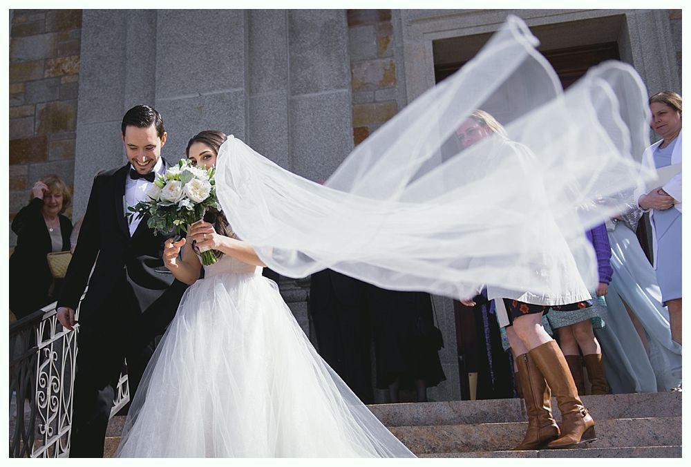 Newlyweds exiting a building, bride's veil billowing. Man in tuxedo, woman in white dress holding flowers.