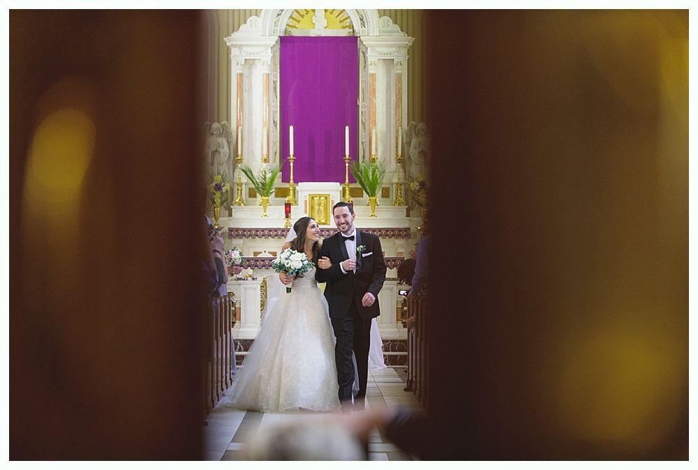 Bride and groom walking down the aisle in a church. Groom wears a tuxedo. Bride holds a bouquet, wearing a white gown.