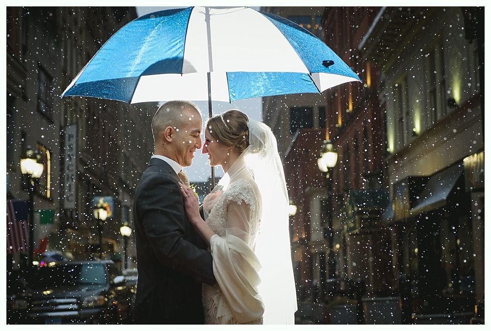Bride and groom under blue and white umbrella, in the rain, in a city street at night.