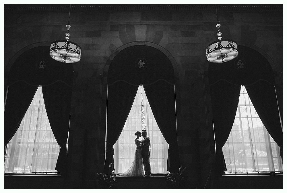Couple embracing in silhouette within arched windows, a grand indoor setting with hanging lamps and dark curtains.