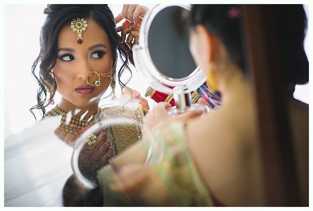 Woman in traditional Indian bridal attire adjusts jewelry in a mirror, indoors.