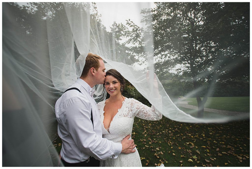 Bride and groom embrace outdoors, veil billowing, groom kissing bride's forehead.
