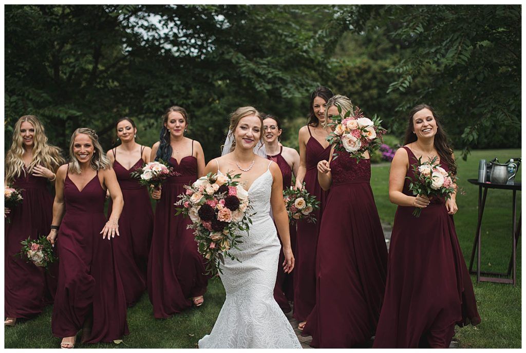Bride smiling, surrounded by bridesmaids in burgundy dresses, holding bouquets outdoors.