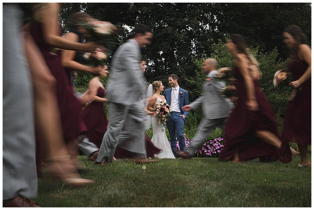 Wedding party surrounds a couple. Bridesmaids in burgundy and groomsmen in gray. Couple smiles amidst motion blur.