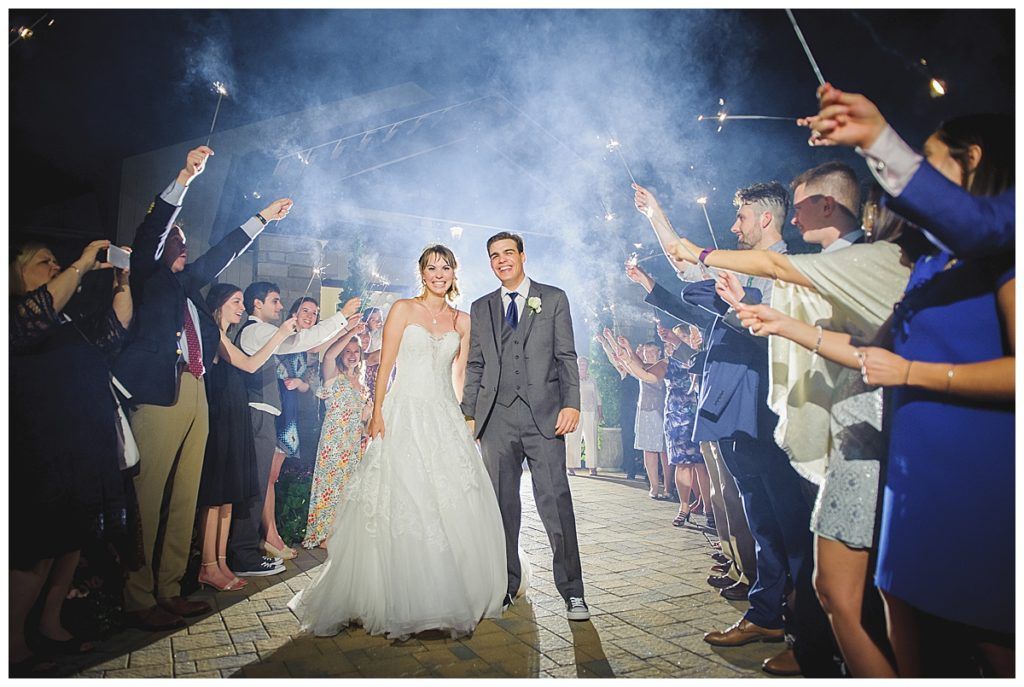 Bride and groom walk between guests holding sparklers after a wedding; celebratory, nighttime.