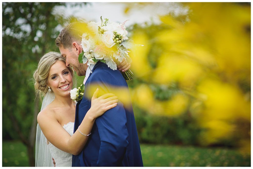 Bride in white dress embraces groom in blue suit outdoors, with yellow flowers in the foreground.